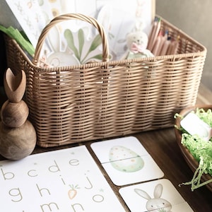 May include: A woven wicker basket filled with Easter-themed items, including a plush bunny, pencils, and decorative paper. A wooden bunny-shaped stacker and alphabet flashcards are in the foreground. A wooden bowl holds a toy carrot and green paper grass.
