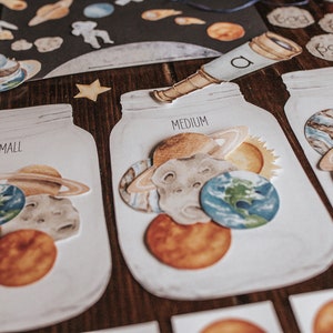 May include: Three white jars with lids, labeled "Small", "Medium", and "Large". Each jar contains different sized paper cutouts of planets, stars, and the sun. The jars are on a wooden surface.