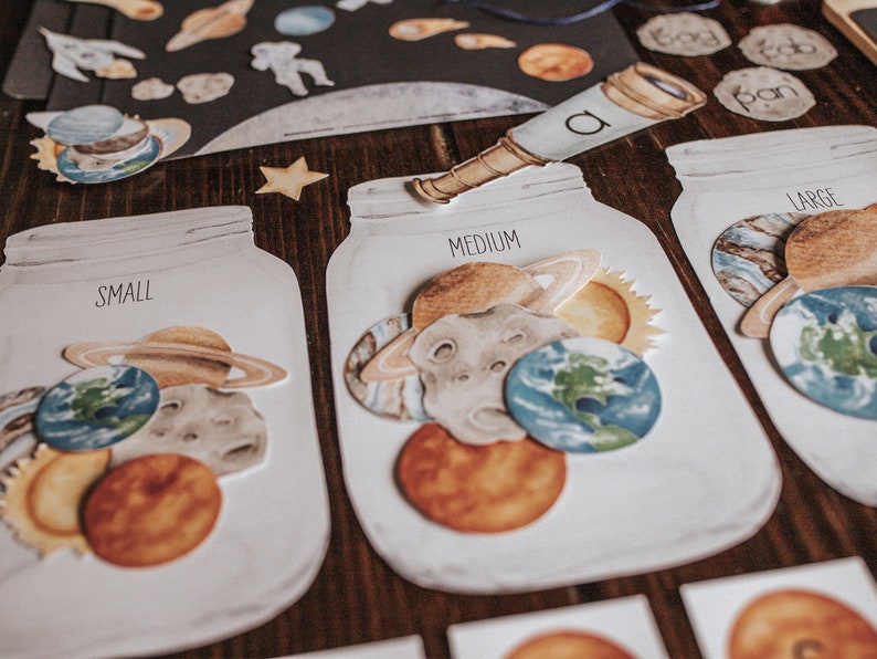 May include: Three white jars with lids, labeled "Small", "Medium", and "Large". Each jar contains different sized paper cutouts of planets, stars, and the sun. The jars are on a wooden surface.