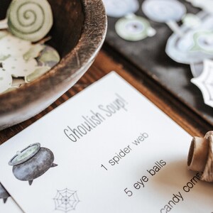 May include: A wooden bowl filled with green and white paper cutouts sits next to a card that reads "Ghoulish Soup!" with a list of ingredients. A cork and other Halloween-themed cutouts are also visible.