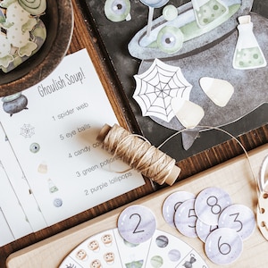 May include: A Halloween-themed craft kit with various paper cutouts, including lollipops, potion bottles, and spiderwebs. A wooden bowl holds additional cutouts. A card reads "Ghoulish Soup!" with a list of ingredients. A wooden board displays number circles.