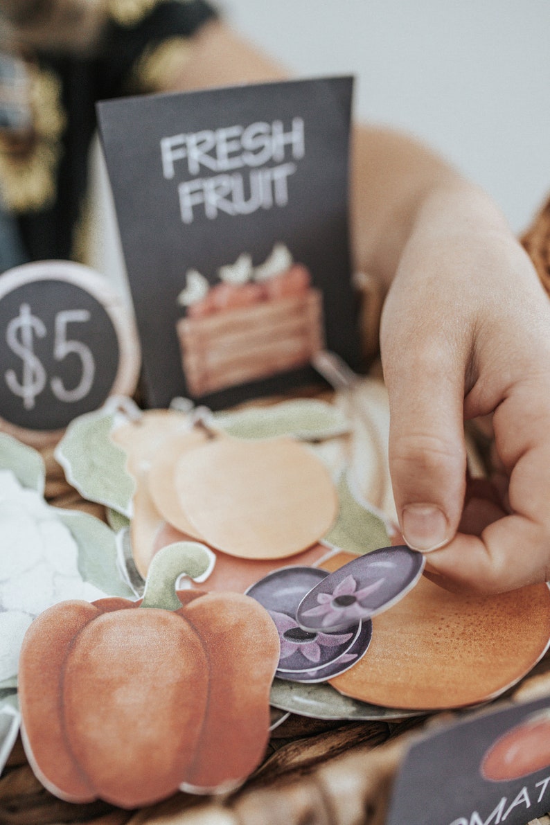 May include: A selection of paper cutouts depicting various fruits and vegetables, such as a red pepper, peaches, and plums. A sign displays "FRESH FRUIT" and another shows the price of &pound;5. A child's hand is holding the cutouts.