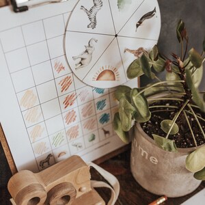 May include: A wooden toy camera with a cream-colored strap sits near a clipboard with a color matching chart and a spinning wheel featuring animal illustrations. A potted plant and a pencil are also visible.