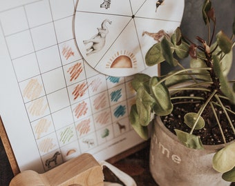 May include: A wooden toy camera with a cream-colored strap sits near a clipboard with a color matching chart and a spinning wheel featuring animal illustrations. A potted plant and a pencil are also visible.