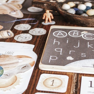 Könnte beinhalten: Lehrmittel und Lernmaterialien auf einer Holzoberfläche. Enthält eine Tafel mit Buchstaben, Planeten-Ausschnitten, Wortkarten, Zahlenkarten und eine kleine Figur. Eine Holzschale enthält bunte Filzkugeln.