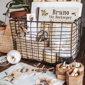 Puede incluir: Una cesta de alambre negra llena de libros, frascos y objetos decorativos con temática de abejas. Los libros incluyen "Bruno the Beekeeper" y "Winnie the Pooh". Una abeja decorativa está unida a la cesta. Otros artículos incluyen recipientes de madera con forma de panal y obras de arte con temática de abejas.