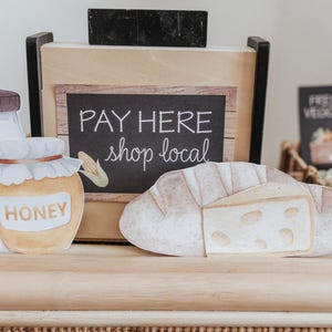 May include: A watercolour illustration of a farmer's market display. Includes a jar of honey, a milk bottle, and a loaf of bread with cheese. A sign reads "PAY HERE shop local". Other signs display "FRESH VEGGIES" and "STRAWBERRY".