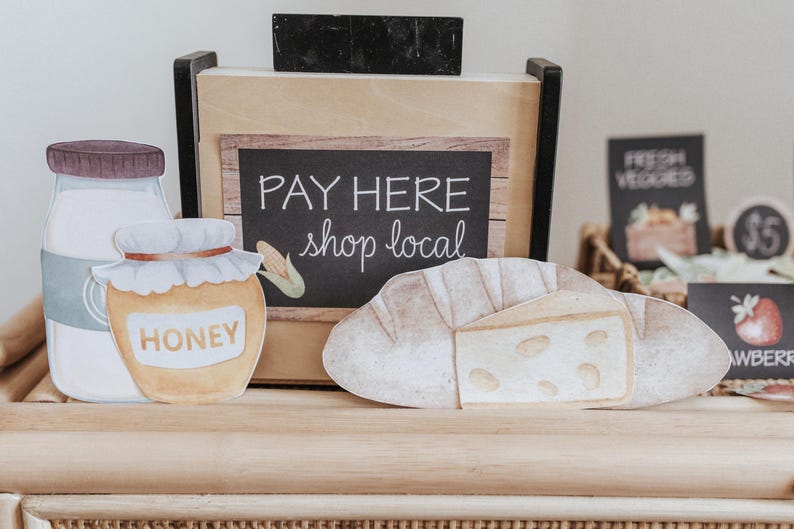 May include: A watercolour illustration of a farmer's market display. Includes a jar of honey, a milk bottle, and a loaf of bread with cheese. A sign reads "PAY HERE shop local". Other signs display "FRESH VEGGIES" and "STRAWBERRY".