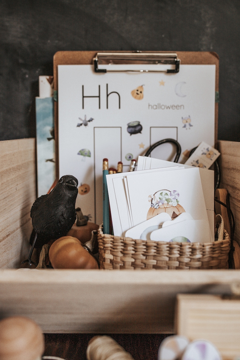 May include: A wooden box filled with Halloween-themed educational materials. Includes a black crow figurine, a clipboard with an alphabet worksheet, and a woven basket containing cards with pumpkin illustrations. Pencils and decorative pumpkins are also present.