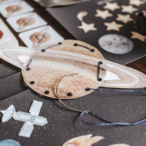 May include: A brown paper craft of Saturn with a white ring and blue thread going through holes around the edge. A needle and thread are on the table.