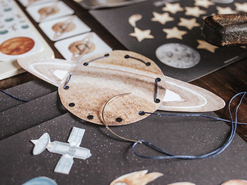 May include: A brown paper craft of Saturn with a white ring and blue thread going through holes around the edge. A needle and thread are on the table.