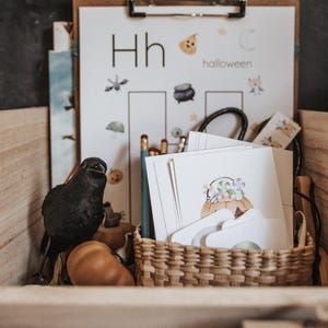 May include: A wooden box contains Halloween-themed educational materials. A black crow figurine sits next to pumpkins. A clipboard displays a worksheet with the letter "H" and the word "halloween". A woven basket holds cards with pumpkin illustrations.