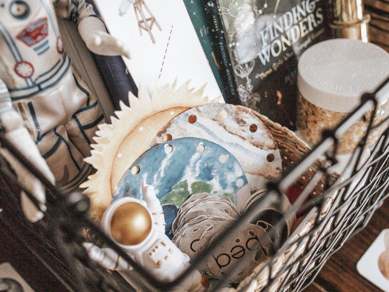 May include: A black wire basket filled with a variety of items, including a toy astronaut, a book titled "Finding Wonders", a wooden sun and moon, and a jar of glitter.