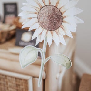 May include: A paper cutout of a sunflower with a brown centre and white petals. The stem and leaves are green. The sunflower is attached to a wooden pole. The background is blurred.