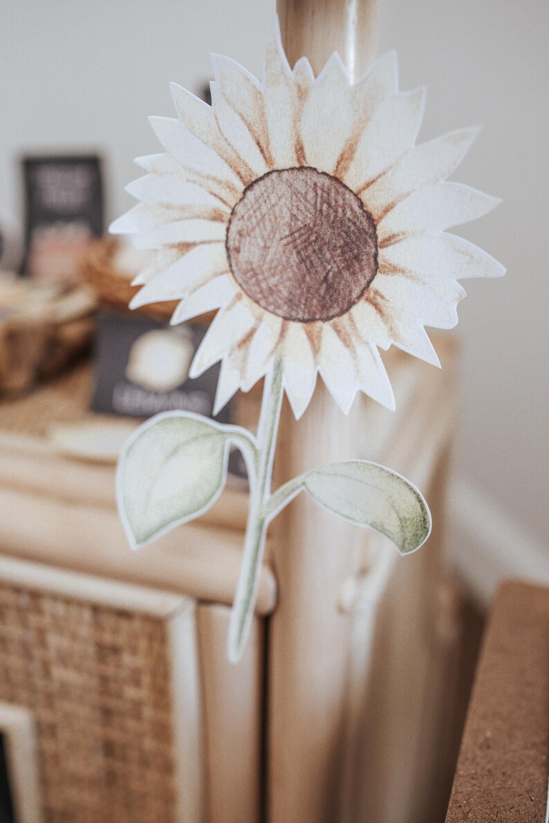 May include: A paper cutout of a sunflower with a brown centre and white petals. The stem and leaves are green. The sunflower is attached to a wooden pole. The background is blurred.