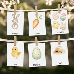 May include: A collection of six cards with Easter-themed illustrations and text. The cards feature a rabbit, carrot, cookies, chick, Easter egg, and a hat. The cards are clipped to a string with wooden clothespins, with a blurred floral background.