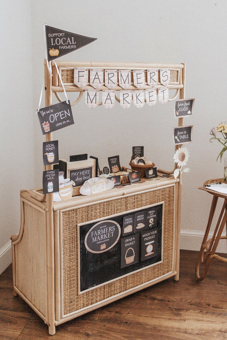 May include: A small farmers market stall constructed from light brown woven material. Signs read "Farmers Market", "Support Local Farmers", and "Open". The stall showcases miniature food items and a chalkboard sign, evoking a rustic charm.