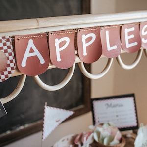 May include: A wooden stand with a red and white checkered banner that says "APPLES" in white letters. The stand has a black sign that says "OPEN come on in..."