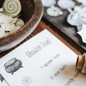 May include: A wooden bowl filled with green and white paper cutouts sits next to a card that reads "Ghoulish Soup!" with a list of ingredients. The card features illustrations of a cauldron, spider web, and eyeball.