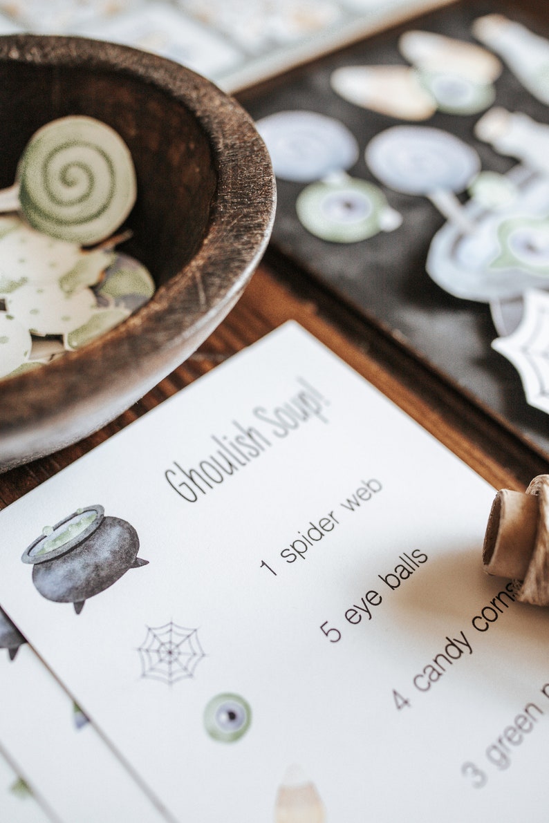 May include: A wooden bowl filled with green and white paper cutouts sits next to a card that reads "Ghoulish Soup!" with a list of ingredients. The card features illustrations of a cauldron, spider web, and eyeball.
