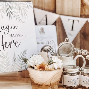 May include: A wooden sign with the text "Magic Happens Here" surrounded by green foliage.  Two glass jars with labels "Beans" and "Seeds" are on a wooden surface with white flowers and a mortar and pestle.