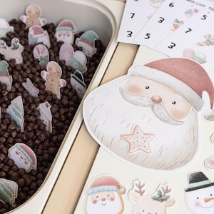 Puede incluir: Un recipiente sensorial con temática navideña con varios recortes de Papá Noel, renos y hombres de jengibre. El recipiente contiene cereales marrones. También hay tarjetas numeradas y un gran recorte de Papá Noel.