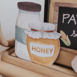 May include: Watercolour illustration of a milk bottle with a brown lid and a honey jar with a white lid and the word "HONEY". The jars are on a wooden surface.