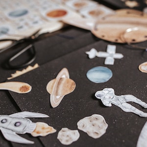 May include: A collection of space-themed stickers on a black background. The stickers include planets, a rocket ship, an astronaut, and a comet.