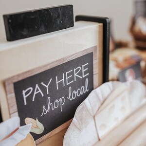 May include: A sign with the words "PAY HERE shop local" in white script. A black rectangular object is on top of the sign. Decorative paper cutouts of food items are in front of the sign. The sign is made of wood.