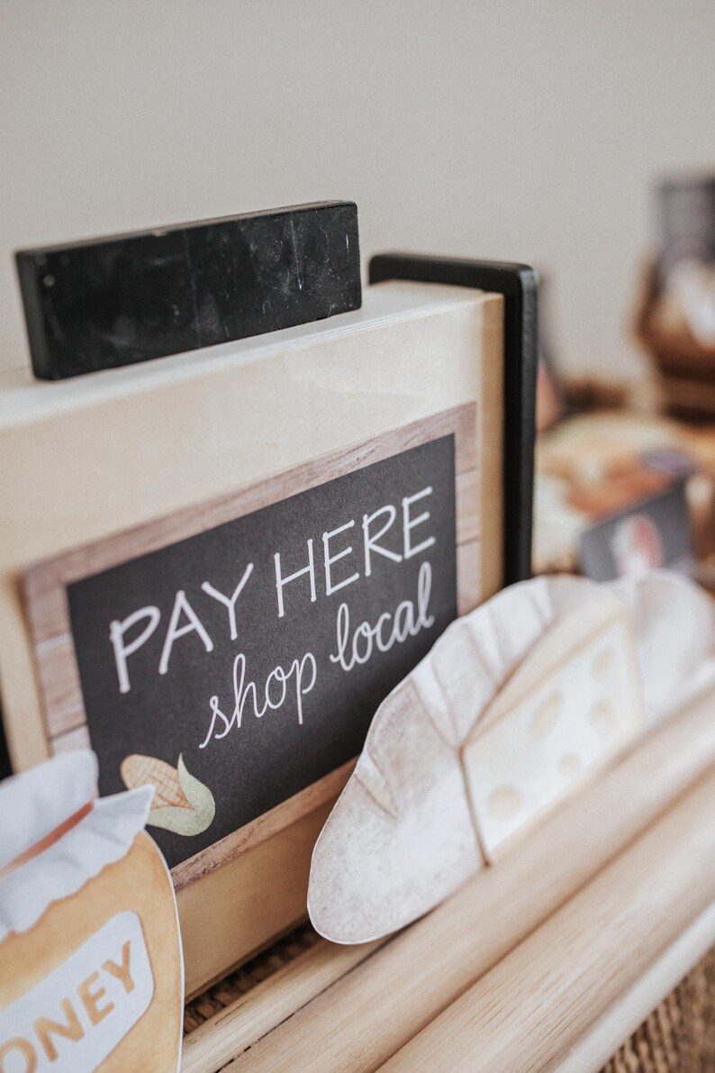 May include: A sign with the words "PAY HERE shop local" in white script. A black rectangular object is on top of the sign. Decorative paper cutouts of food items are in front of the sign. The sign is made of wood.