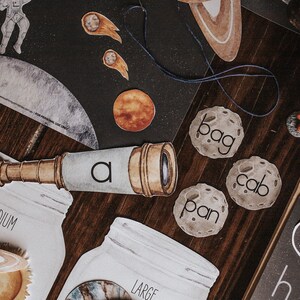 May include: A set of space-themed educational materials for children, including a telescope, a moon with the word "bag" on it, a moon with the word "cab" on it, a moon with the word "pan" on it, and a chalkboard with the letters "A" and "hi" on it.