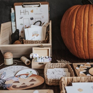May include: A wooden box and baskets filled with Halloween-themed educational materials. Includes a large orange pumpkin, a clipboard with the word "Halloween", and various cutouts of pumpkins, candy corn, and spiders. A small wooden box contains decorative cutouts.