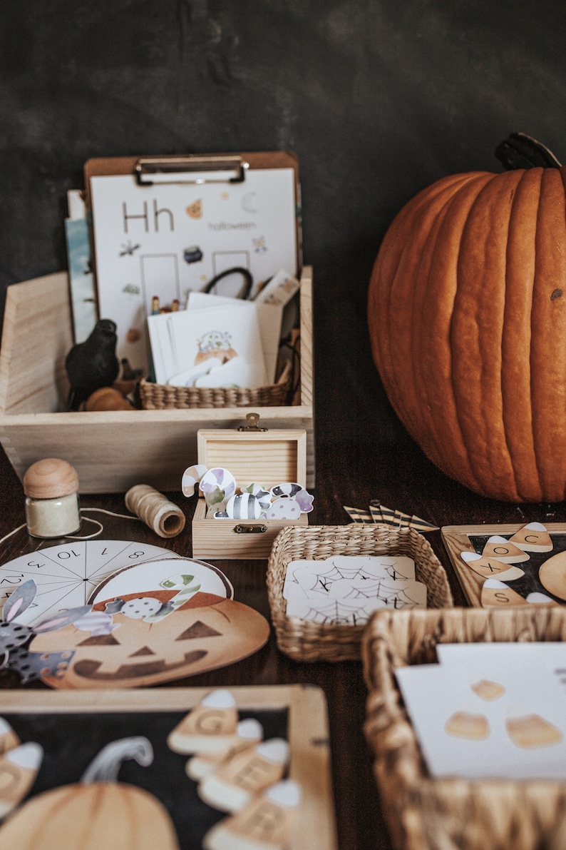 May include: A wooden box and baskets filled with Halloween-themed educational materials. Includes a large orange pumpkin, a clipboard with the word "Halloween", and various cutouts of pumpkins, candy corn, and spiders. A small wooden box contains decorative cutouts.