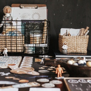 May include: A black wire basket filled with space-themed toys and learning materials, including an astronaut figurine, a book titled "Finding Wonders", and a globe. The basket is on a table with other space-themed toys, including a wooden bowl filled with colorful balls, a black chalkboard with white circles, and a set of cards.
