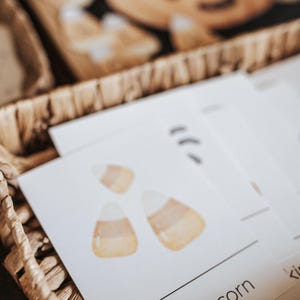 May include: A woven basket holds white cards with watercolor candy corn illustrations. The words "candy corn" are printed in a black, sans-serif font. A wooden pumpkin decoration is visible in the background.