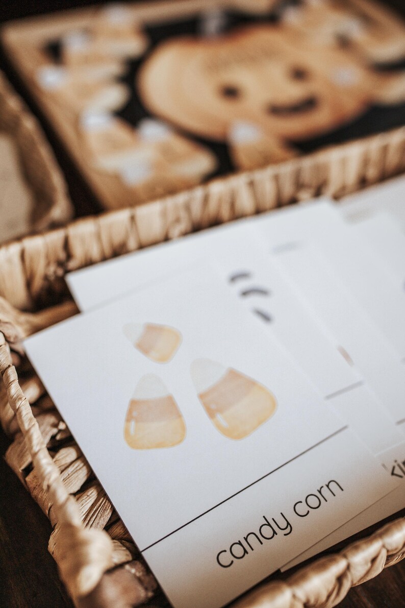 May include: A woven basket holds white cards with watercolor candy corn illustrations. The words "candy corn" are printed in a black, sans-serif font. A wooden pumpkin decoration is visible in the background.