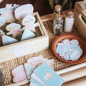 May include: A wooden box filled with colourful paper cutouts of gingerbread people, cupcakes, and candy canes. The box is sitting on a woven surface with a small bowl of candy sprinkles and two glass jars with lids.