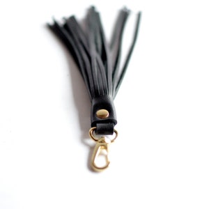May include: A black leather tassel with a gold-colored clasp and rivet. The tassel is made of multiple strips of black leather, and the clasp is a lobster claw style. The background is white.