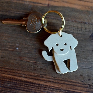 May include: A light gray leather dog-shaped keychain with a gold-colored key ring. The dog has a smiling face and is attached to a gold-colored key. The background is a dark wooden surface.