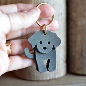 May include: A gray leather dog-shaped keychain with a gold-colored ring. The dog has a layered design with a small tail and cut-out legs. The keychain is held up by a hand.