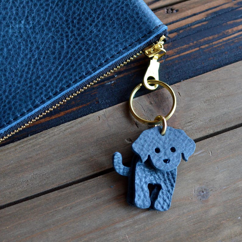 May include: A blue leather dog keychain with a gold-colored ring and clasp. The keychain is next to a blue leather wallet with a gold zipper. The background is a wooden surface.