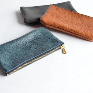 May include: Three leather zipper pouches in different colors: blue, brown, and gray. The pouches are made of textured leather and have a gold zipper.
