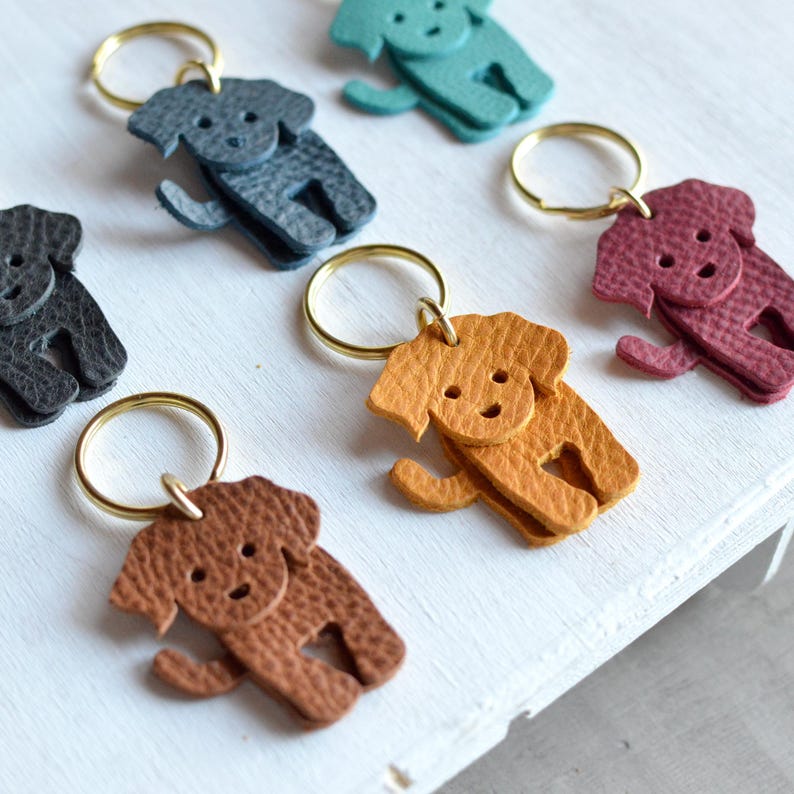 May include: A collection of leather dog-shaped keychains in various colors, including brown, gray, teal, and burgundy. Each keychain features a gold-colored ring for attaching keys. The keychains are arranged on a white surface.