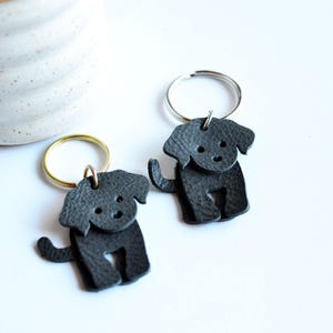 May include: Two dark gray leather dog-shaped keychains. Each keychain features a small, stylized dog with a round head, floppy ears, and a curled tail. The keychains have metal rings for attaching keys.