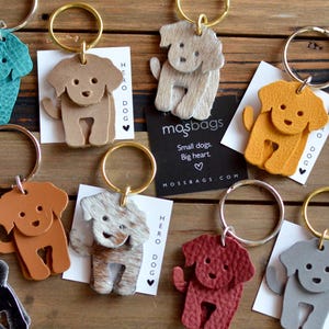 May include: A collection of dog-shaped keychains in various colors, including tan, brown, and red. Each keychain features a gold-colored ring and a small tag with the text "HERO DOG" and "mosbags.com". The keychains are made of leather or a similar material.