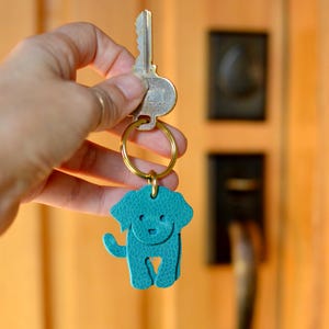 May include: A teal leather dog-shaped keychain with a gold-colored ring and key. The dog has a smiling face and a tail. The keychain is hanging from a key. The background is a wooden door.