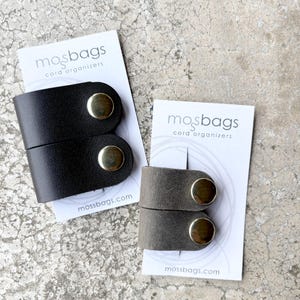 May include: Two sets of cord organizers on white display cards labeled mosbags cord organizers. One set is dark brown leather with gold-tone metal snaps, and the other is a smaller gray leather set with matching gold-tone metal snaps.