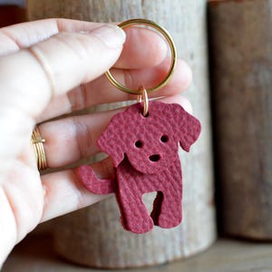 May include: A burgundy leather dog-shaped keychain with a gold-colored ring. The dog has a textured surface with cut-out eyes and a smiling mouth. The keychain is held up against a blurred background.