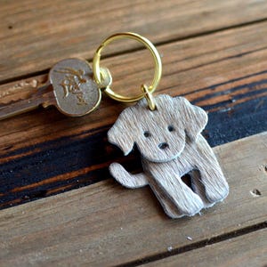 May include: A gold-toned metal keyring attached to a metallic key and a small, flat, dog-shaped charm. The charm is covered in a textured, light brown, hair-like material, featuring two small circular indentations for eyes and a tiny dot for a nose.