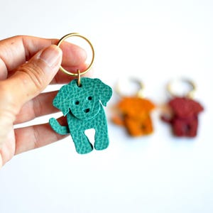 May include: A teal leather dog-shaped keychain with a gold-colored ring. The dog has a textured surface and a simple face. Two other keychains, orange and red, are slightly out of focus in the background.
